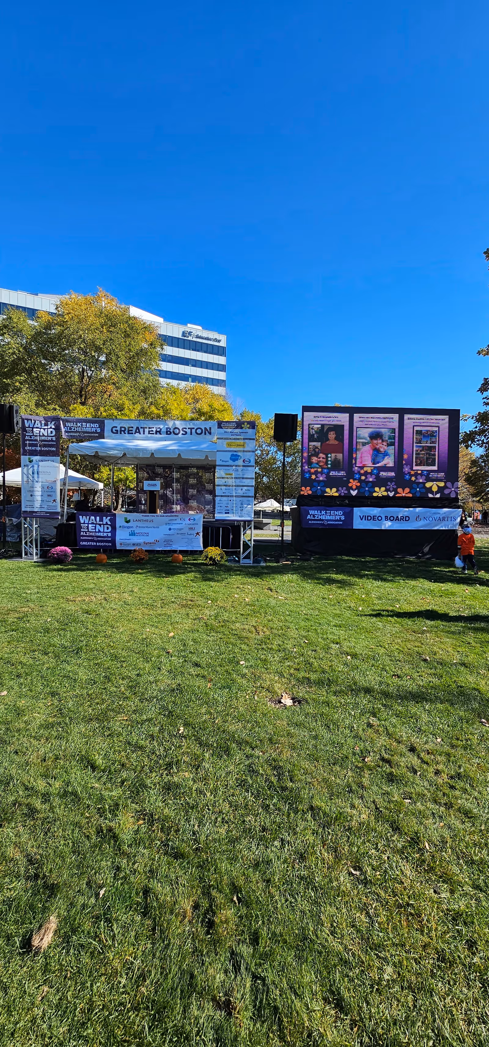 Outdoor park setup with a stage, banners, tents, and a grassy field under blue sky