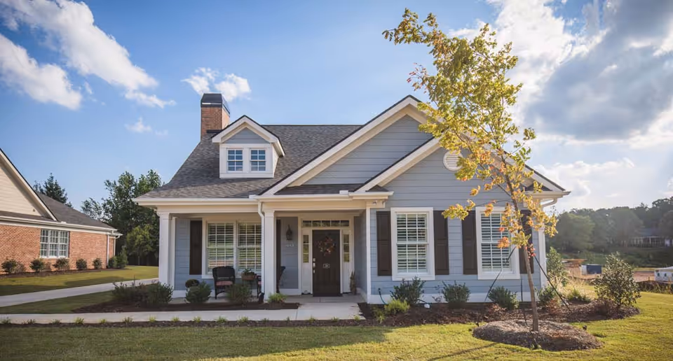 Front exterior view of a single-story house with gray siding, white trim, and a dark roof. The house has a small porch with two chairs and a wreath on the front door. There is a young tree and landscaped bushes in the front yard under a partly cloudy sky.