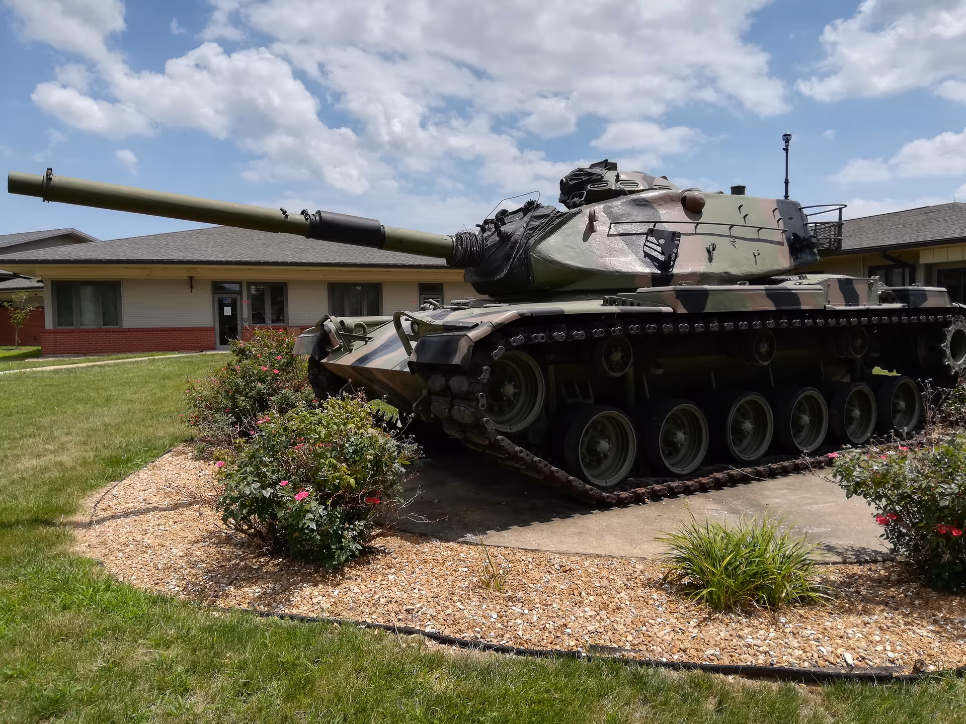 A camouflaged military tank is displayed in a garden area in front of a building.
