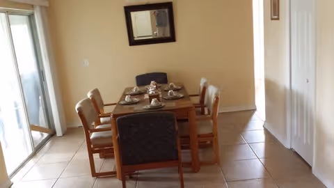 A dining area with a rectangular wooden table set for six people. The table has six chairs, with two chairs at each long side and one chair at each end. The table is set with plates, cups, and napkins. There is a large window or sliding glass door to the left, allowing natural light into the room. A mirror is mounted on the beige wall above the table, and the floor is tiled.