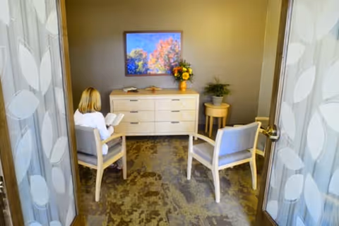A small living room with two chairs, a dresser, TV, and a resident reading.
