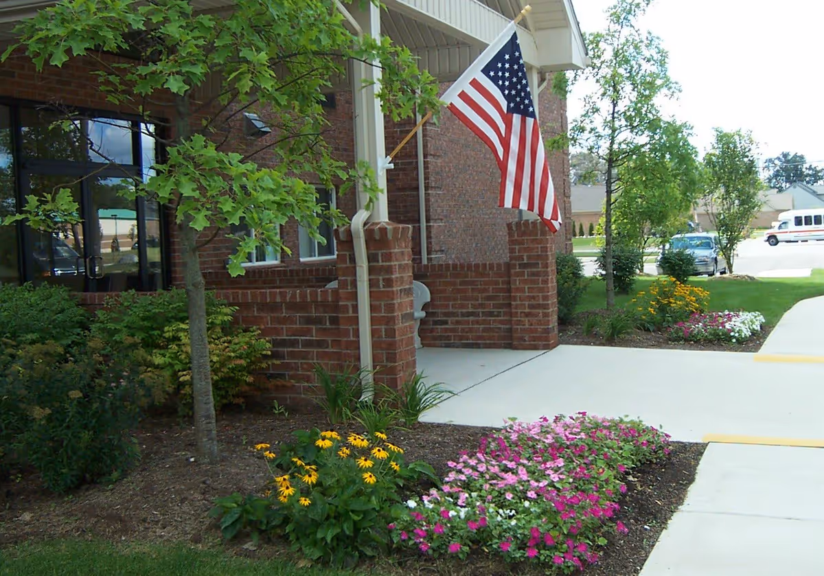 Brick building entrance with an American flag, sidewalk and flower beds in front.