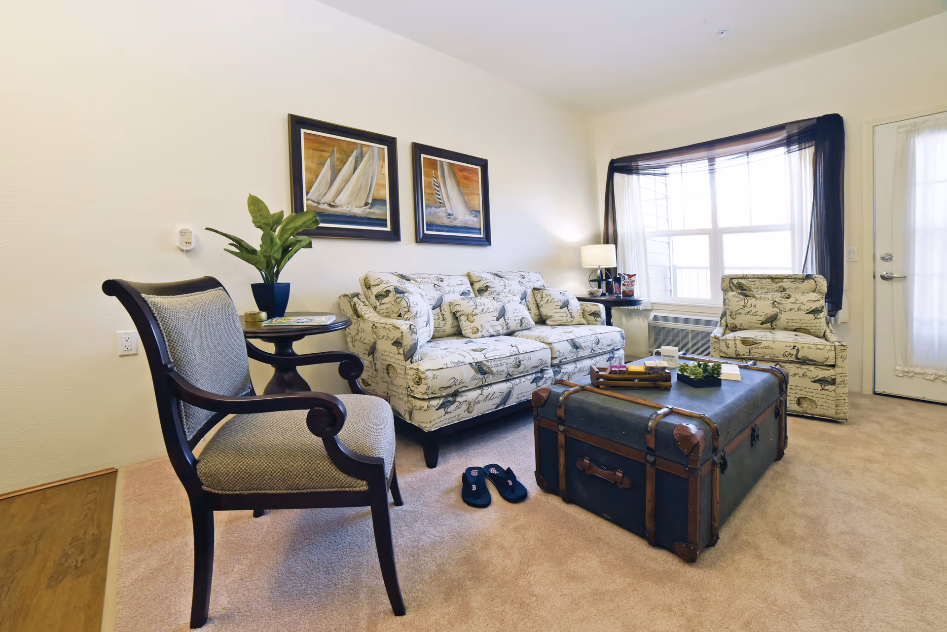 A cozy living room with a patterned sofa and matching armchair, a wooden armchair with cushioned seat, a round side table with a potted plant, and a large vintage trunk used as a coffee table. The room has beige carpet, two framed sailboat paintings on the wall, a window with sheer curtains, and a door with a glass panel.