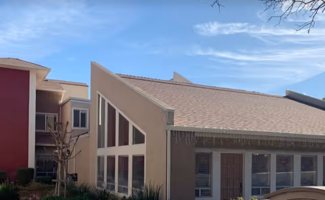 Exterior view of a senior living facility building with a slanted roof and large triangular windows. The building is beige with a red section on the left side. There are some small bushes and a leafless tree in front of the building under a clear blue sky.