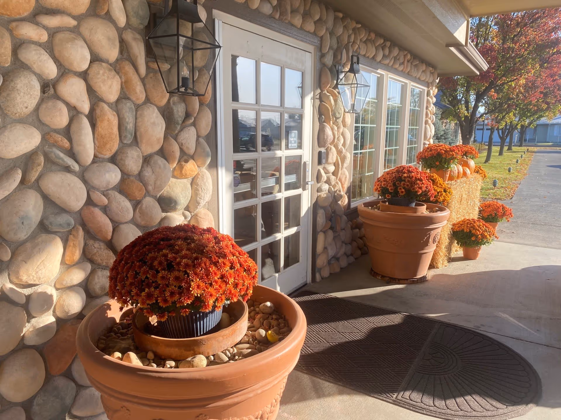 Entrance area of a building with a stone exterior wall, a white door with glass panes, and large windows. There are several large terracotta pots filled with orange and red flowers, along with hay bales decorated with pumpkins and flowers. The scene is sunlit with a paved walkway and trees with autumn foliage in the background.