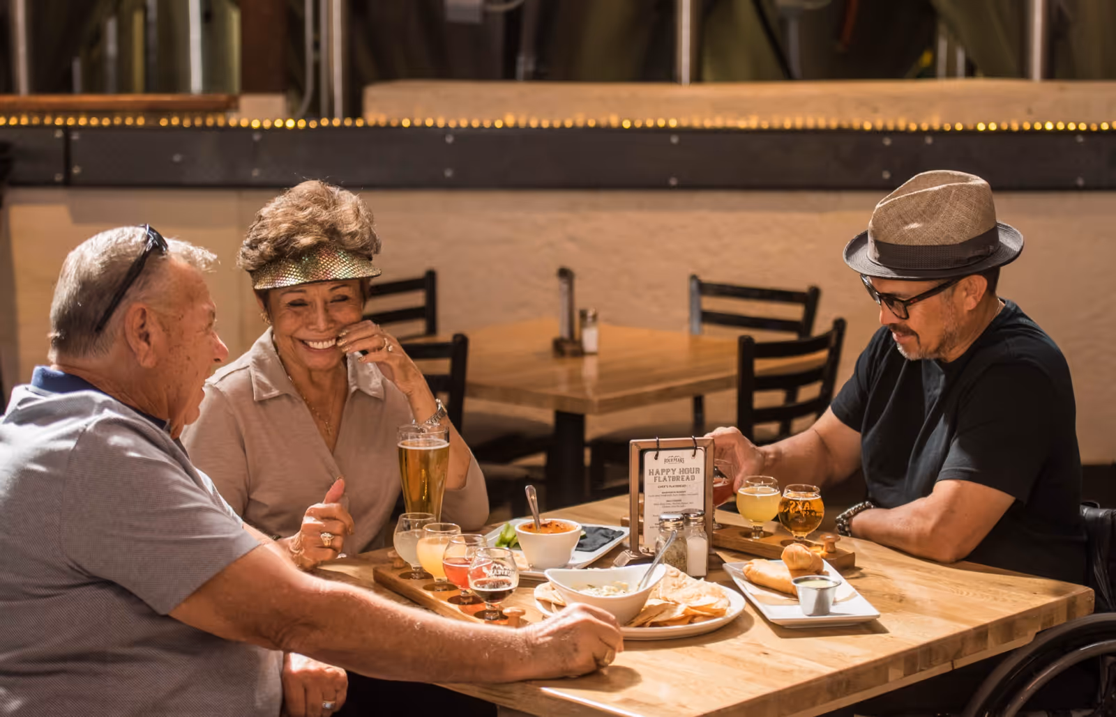 Three older adults seated at a wooden table enjoying drinks and appetizers in a restaurant-style dining area.