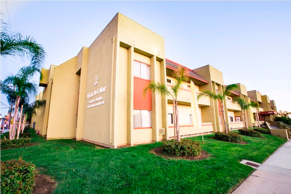 Exterior of the Vista Del Mar Senior Living building with beige-and-red facade, palm trees, and a manicured lawn.