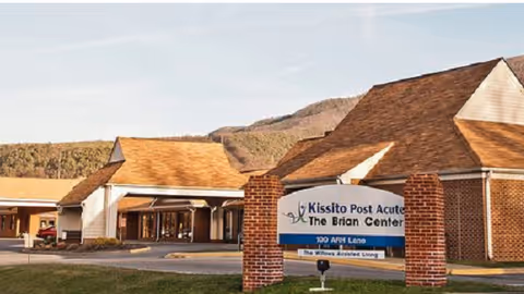 Exterior view of Kissito Post Acute - The Brian Center Alleghany building with a sign in front displaying the facility name and address. The building has a brick and white exterior with a peaked roof, set against a backdrop of hills and a clear sky.