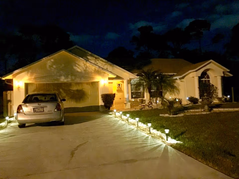 Illuminated single-story house front at night with a car in the driveway and pathway lights leading to the entrance.