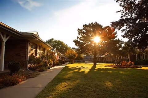 Sunlit senior living campus with brick buildings, a grassy lawn, and a paved walkway.