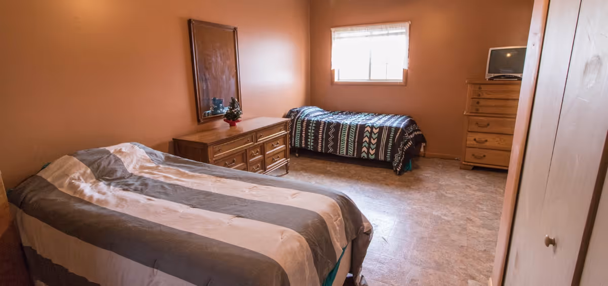 A bedroom with two beds, one with a striped gray and white comforter and the other with a patterned blue and black comforter. There is a wooden dresser with a mirror above it and a small decorative plant on top. A window with blinds is on the far wall, and a wooden chest of drawers with a small TV on top is in the corner. The walls are painted a warm brown color and the floor has a light-colored tile.
