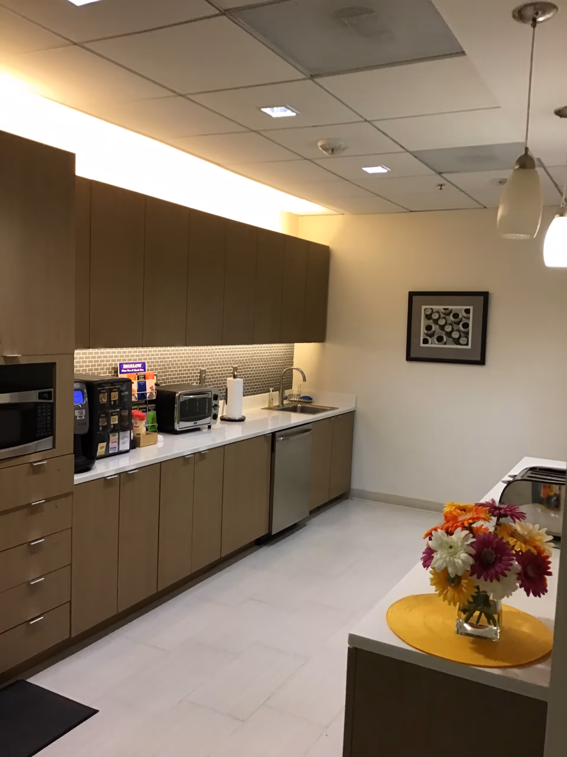 Long modern kitchen with brown cabinets, appliances, sink, and a vase of flowers on the counter.