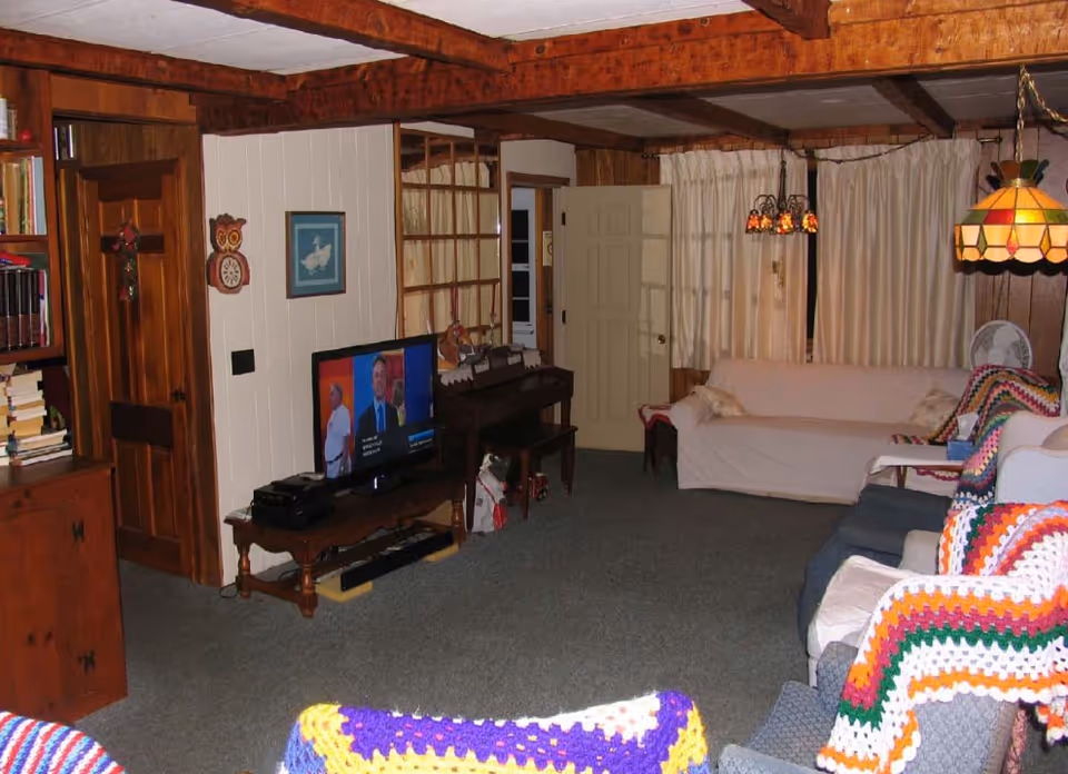 A cozy living room with wooden beams on the ceiling and wood-paneled walls. The room features a television on a wooden stand, a piano, a white couch, and several armchairs covered with colorful crocheted blankets. There are cream-colored curtains covering the windows and two hanging stained glass lamps providing warm lighting.