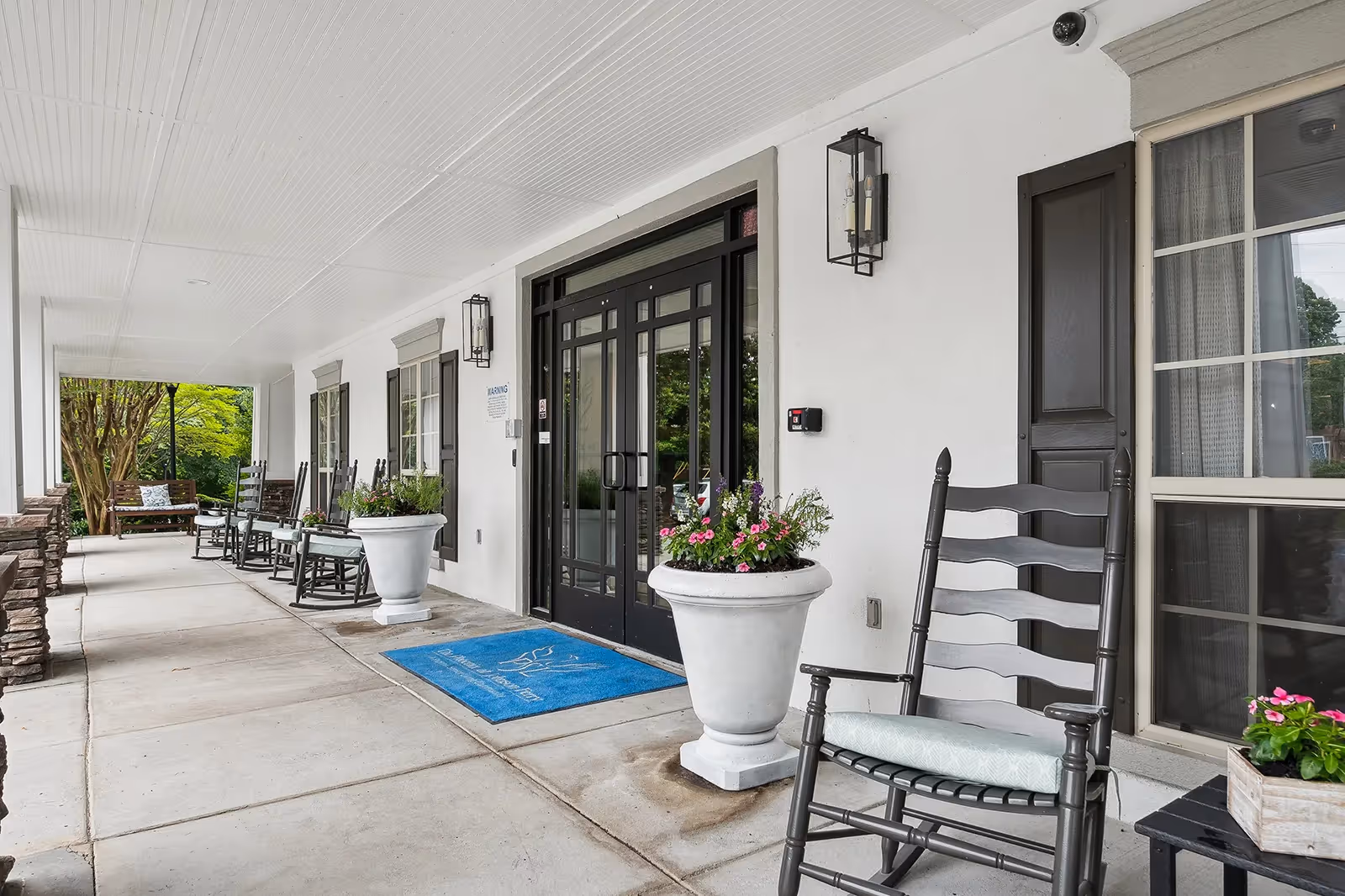 Covered front porch entrance with black double doors, potted flowers, a blue welcome mat, and several rocking chairs along a concrete walkway.