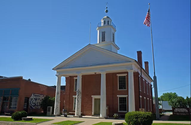 Red brick classical-style building with a white columned portico, a cupola on the roof, and an American flag on a pole against a clear blue sky.
