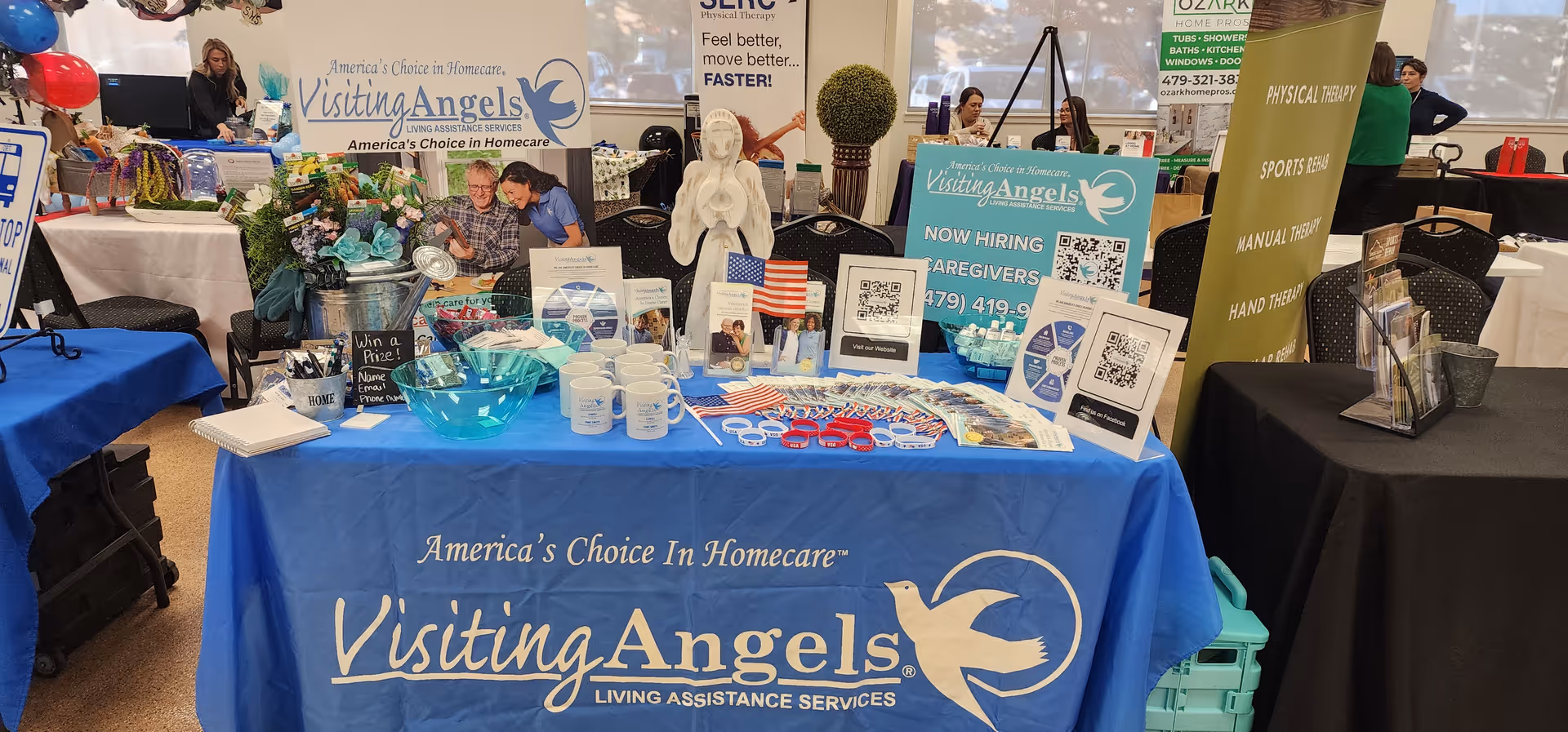Indoor Visiting Angels booth at Fort Smith expo with tables, banners, and staff.