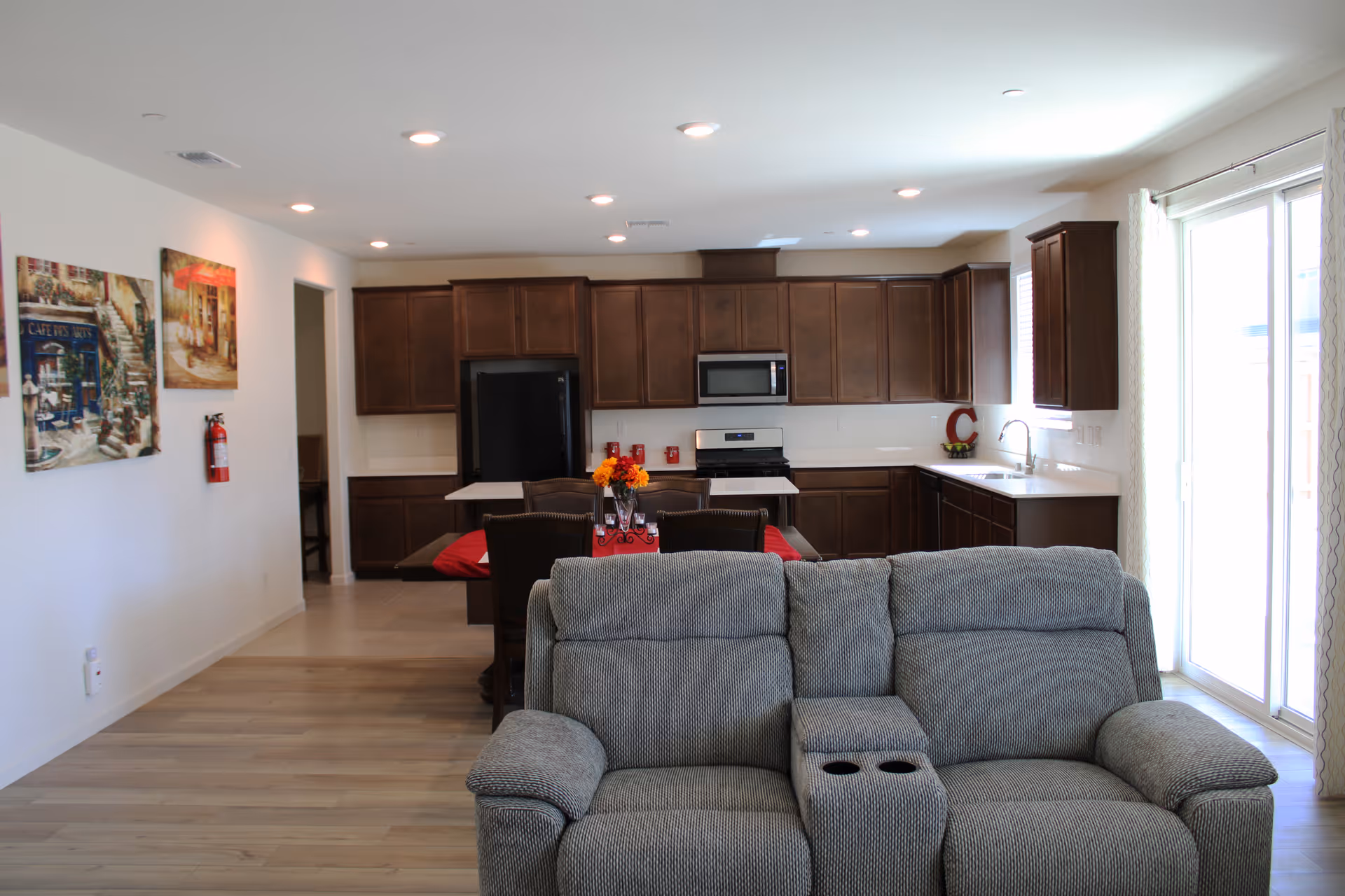 Interior view of a senior living facility showing a living room area with a gray reclining sofa facing away from the camera, and an open kitchen with dark wooden cabinets, a black refrigerator, a microwave, and a stove. There is a dining table with chairs and a red table runner with a flower centerpiece. The room has light-colored walls, wooden flooring, and large sliding glass doors with curtains letting in natural light.