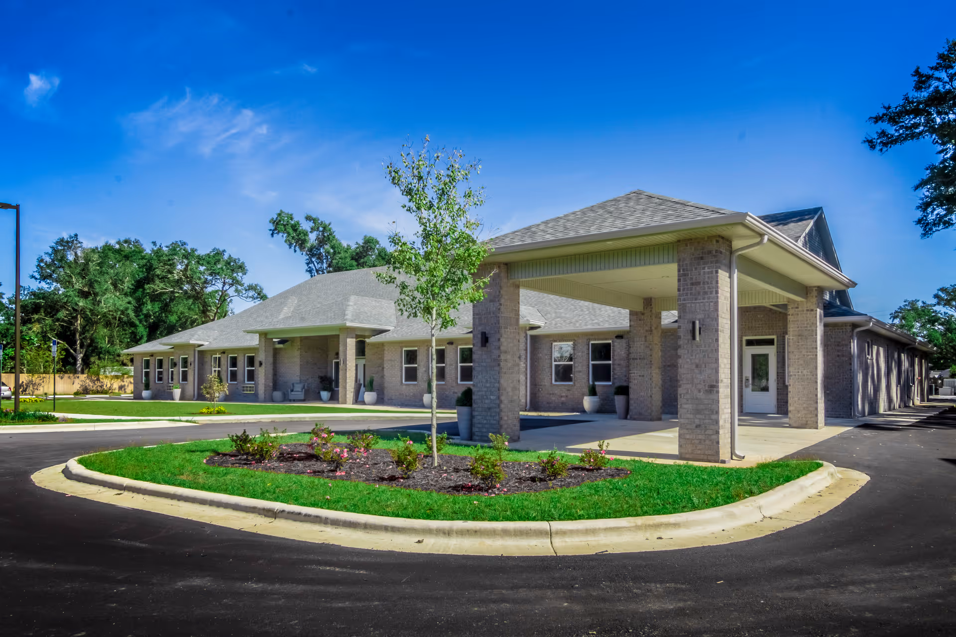 Exterior view of Shelton Manor Independent and Assisted Living facility showing a single-story brick building with a covered entrance, surrounded by a circular driveway and landscaped greenery under a clear blue sky.