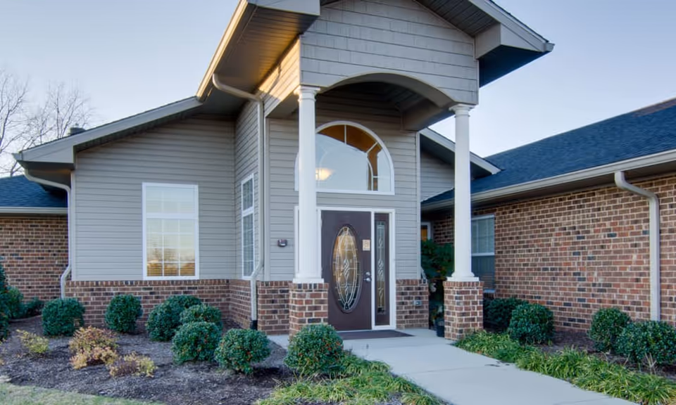 Front entrance of a single-story brick and siding building with a covered portico, decorative glass door, walkway, and landscaped shrubs.