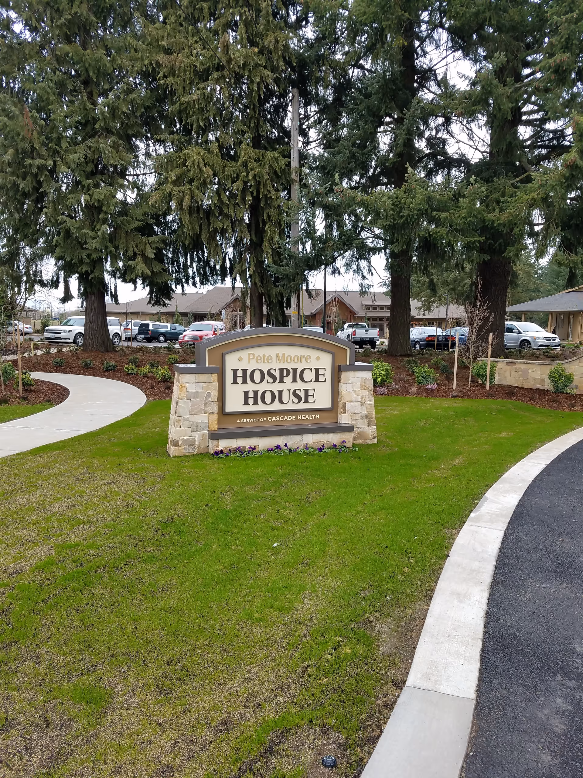 Exterior entrance sign reads Pete Moore Hospice House amid lawn, trees, and parking.