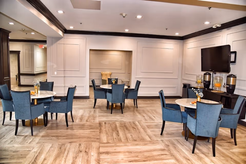 A well-lit dining area with several round tables, each surrounded by blue upholstered chairs. The room features light wood flooring, white paneled walls with dark trim, and a flat-screen TV mounted on the wall. There are beverage dispensers on a side table beneath the TV, and small floral centerpieces on each dining table.