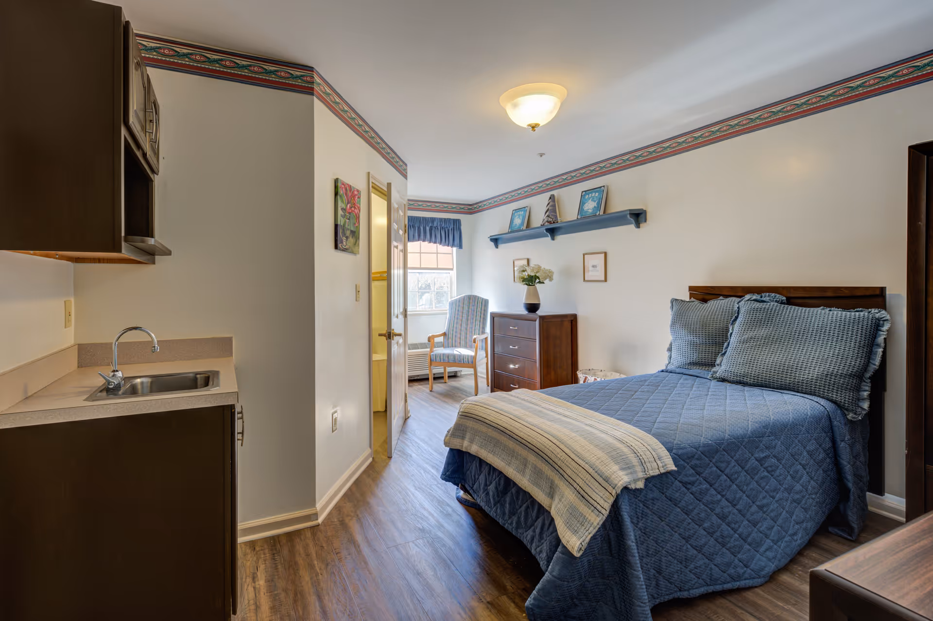 Cozy bedroom with blue quilt, dresser, kitchenette area, and seating by window.