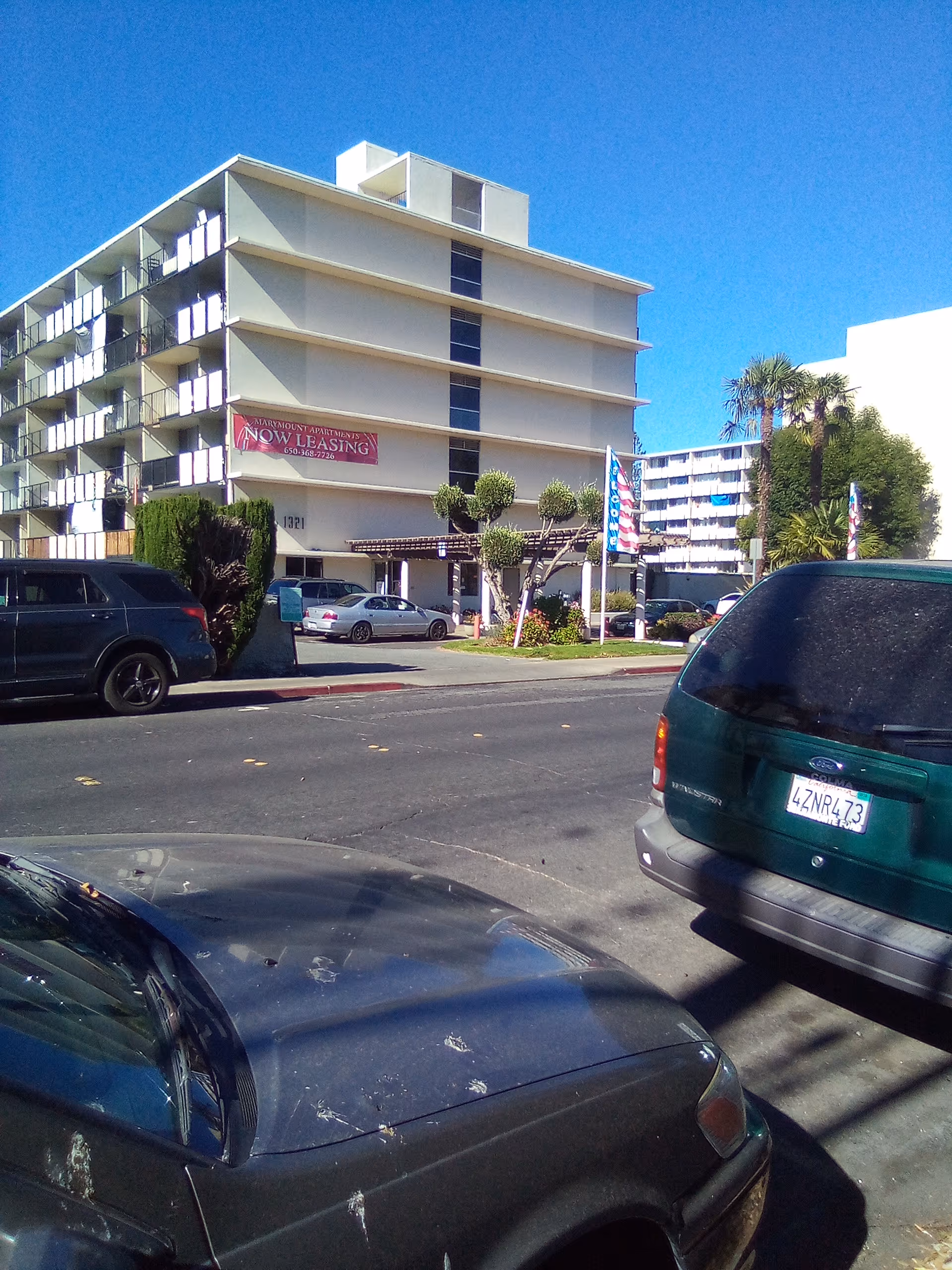 A multi-story apartment-style building with balconies, parked cars, and a 'Now Leasing' sign.