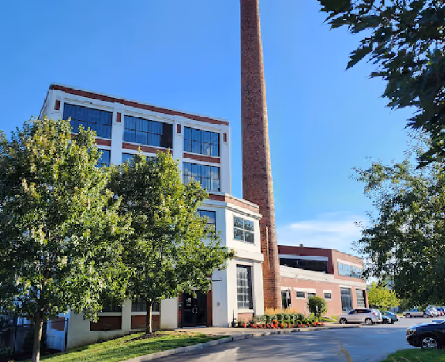 Exterior view of a multi-story brick building with a tall chimney, trees, and parked cars.