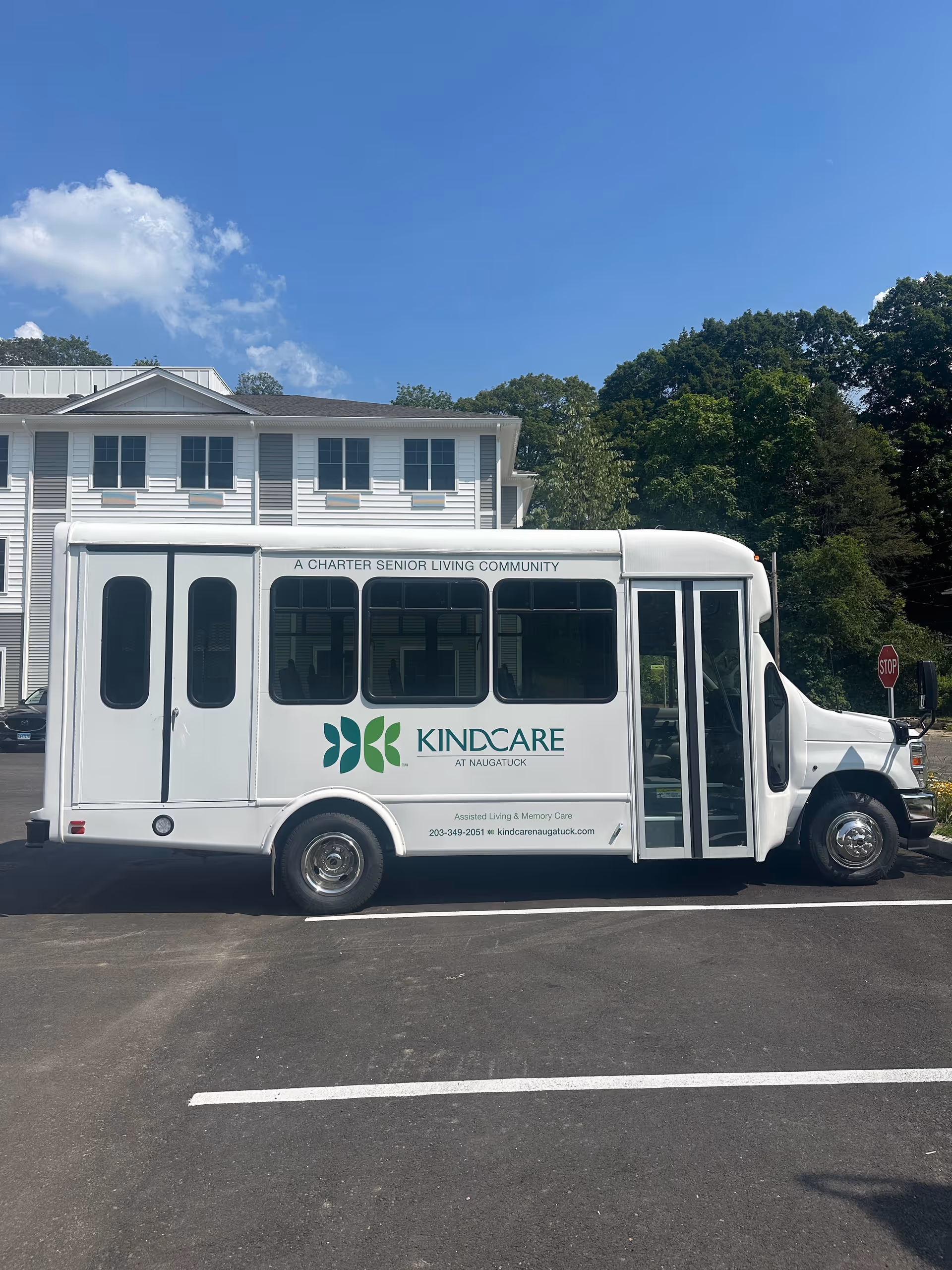 White KindCare Naugatuck shuttle bus parked in a lot with a building and trees behind