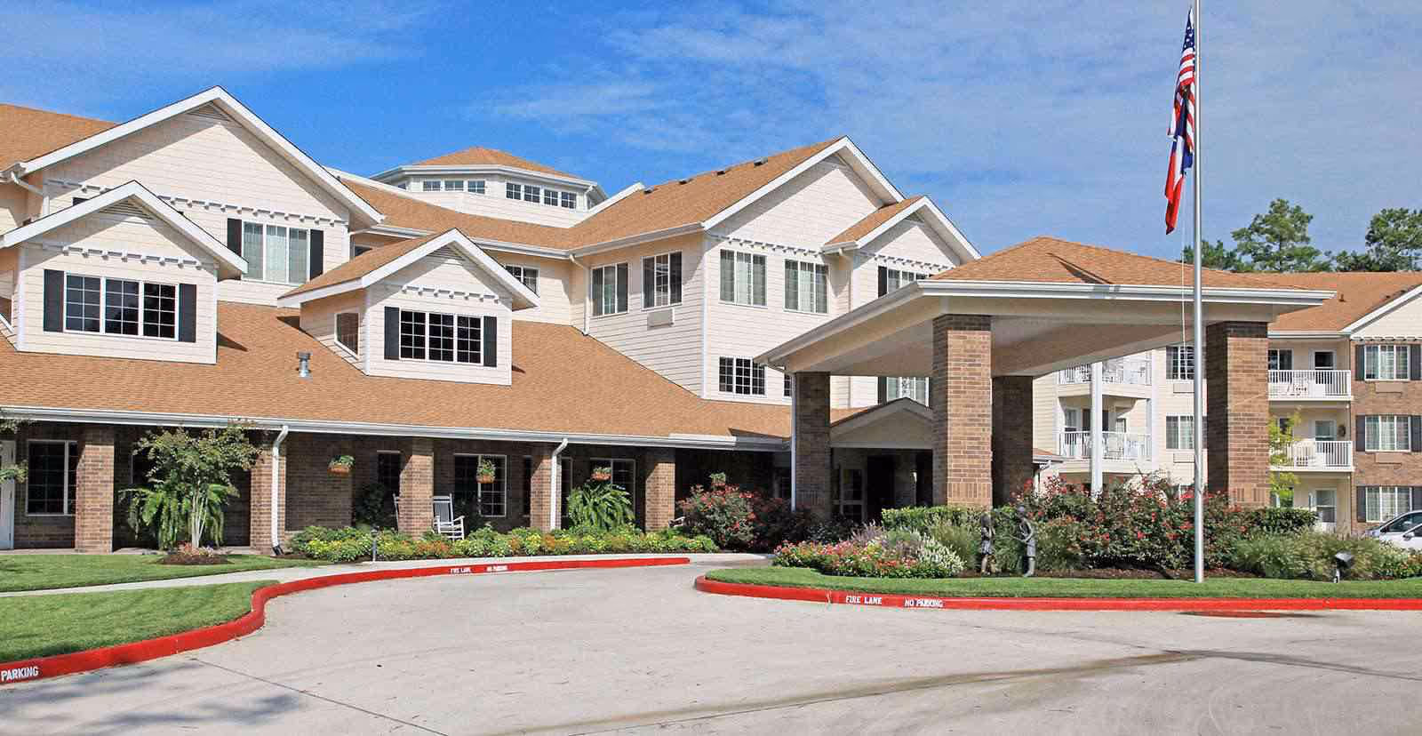 Front entrance of a multi-story senior living building with a covered porte-cochere, landscaped driveway, and a flagpole.