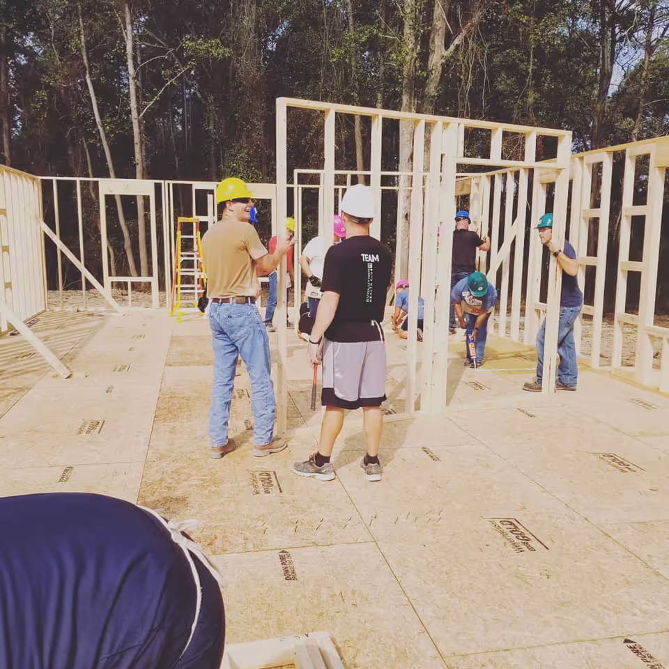 Construction crew wearing hard hats work on wooden wall frames outdoors.