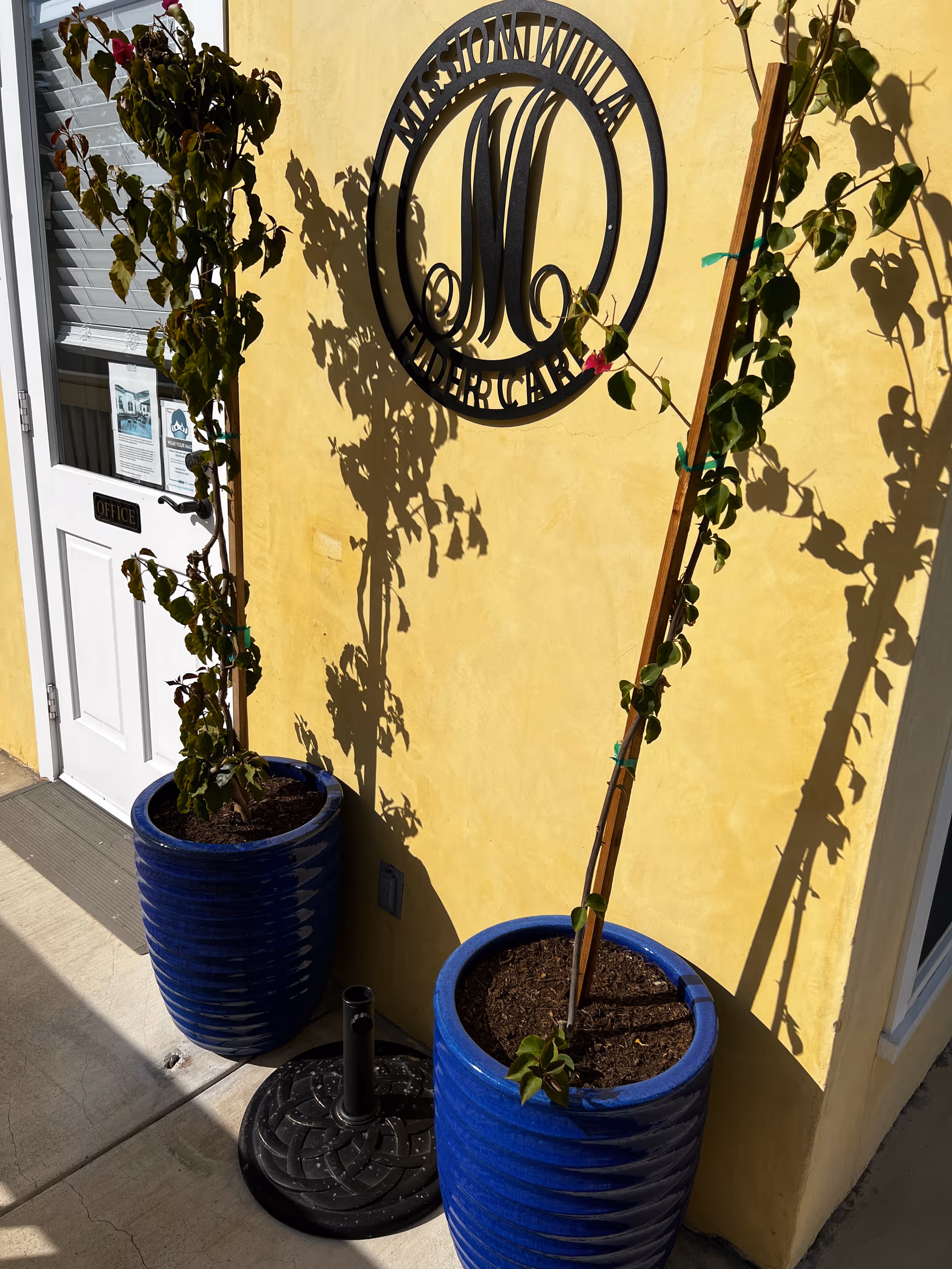 Exterior yellow wall with Mission Villa circular logo, two blue planters, and a door.