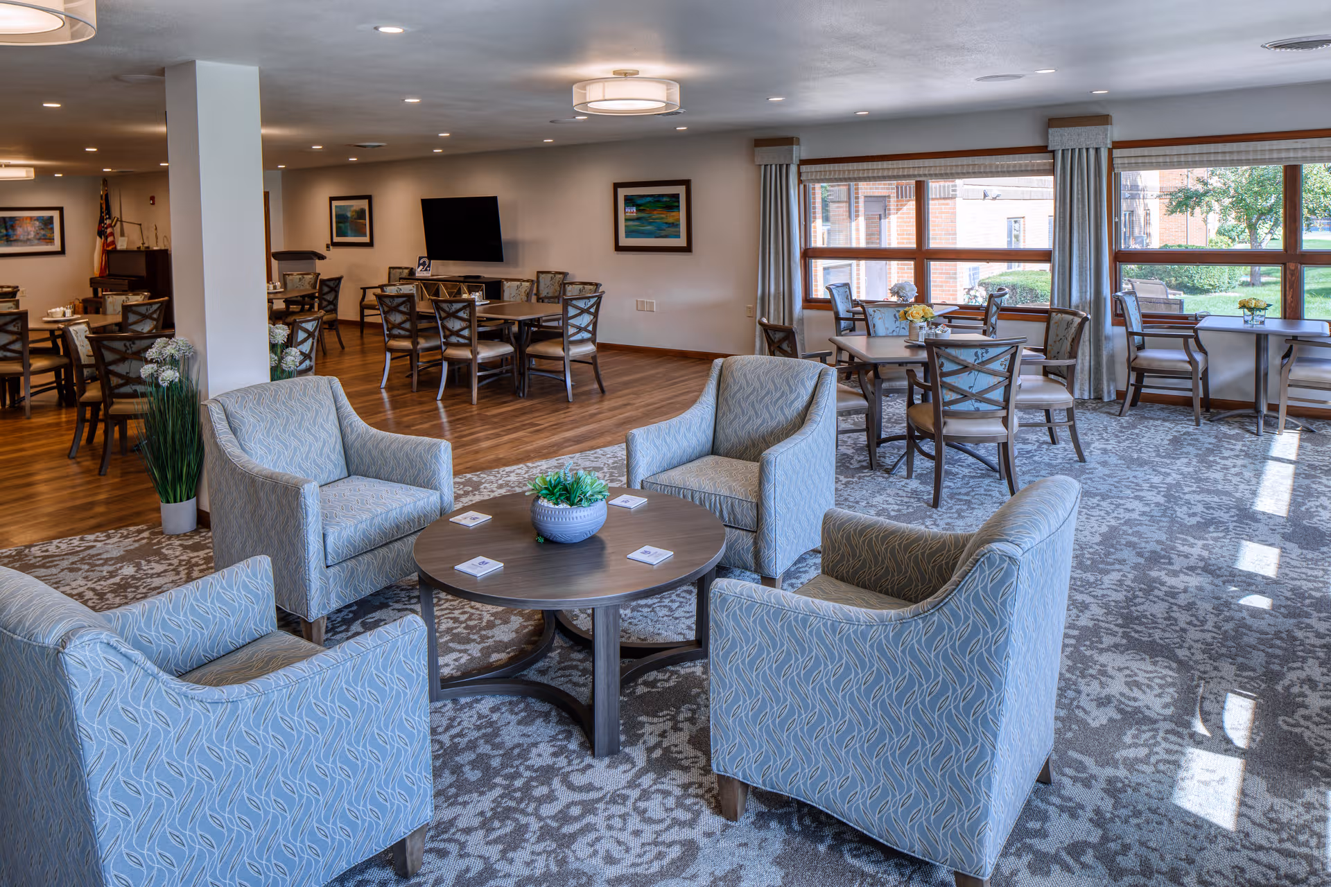 Common lounge area with blue patterned chairs, round table, and large windows.