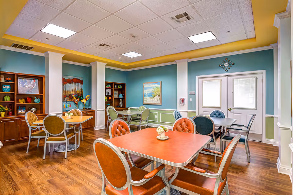 Bright, colorful senior living dining room with multiple tables, chairs, and wall art.