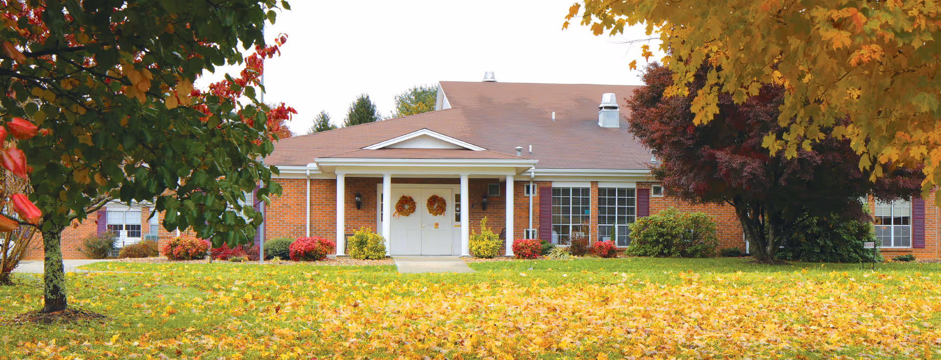 Brick senior living facility with a front entrance, autumn trees, and fallen leaves.