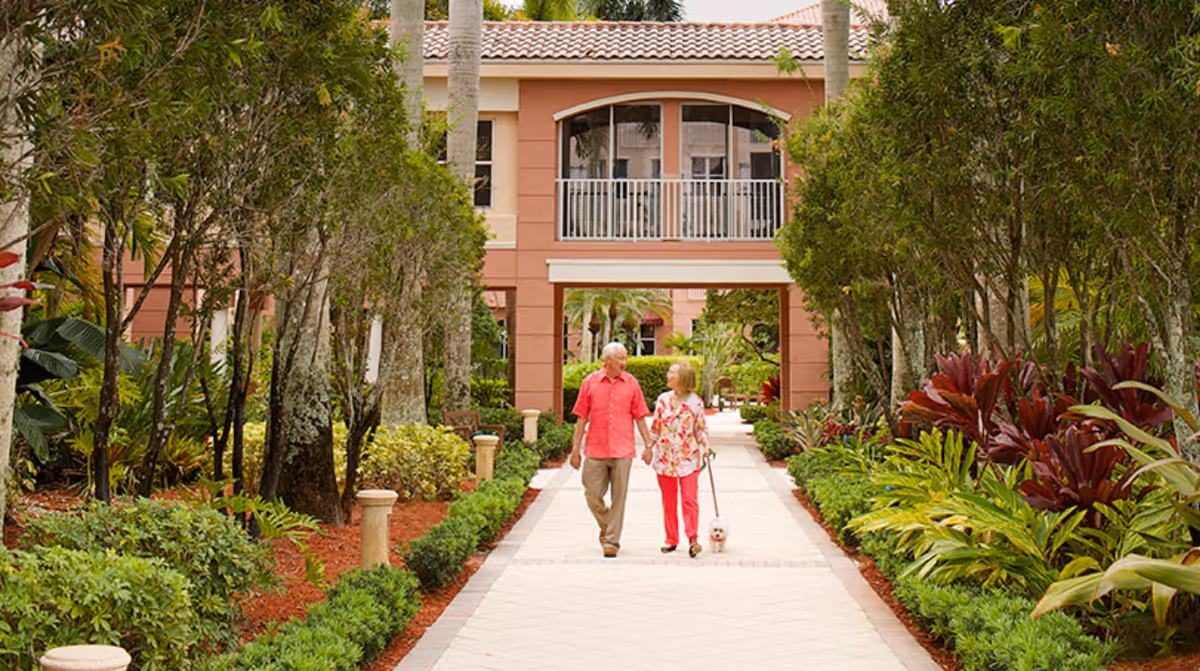 An older couple walking a small dog along a landscaped pathway toward a peach-colored senior living building.