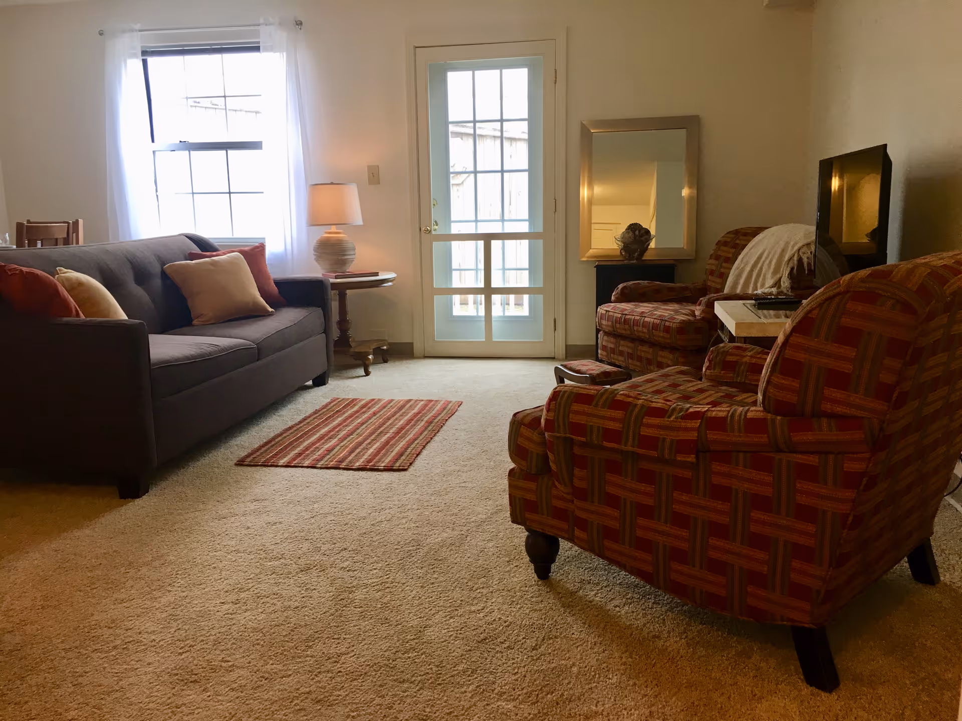 Cozy living room with sofa, plaid chairs, rug, lamp, TV, and glass-door entry.