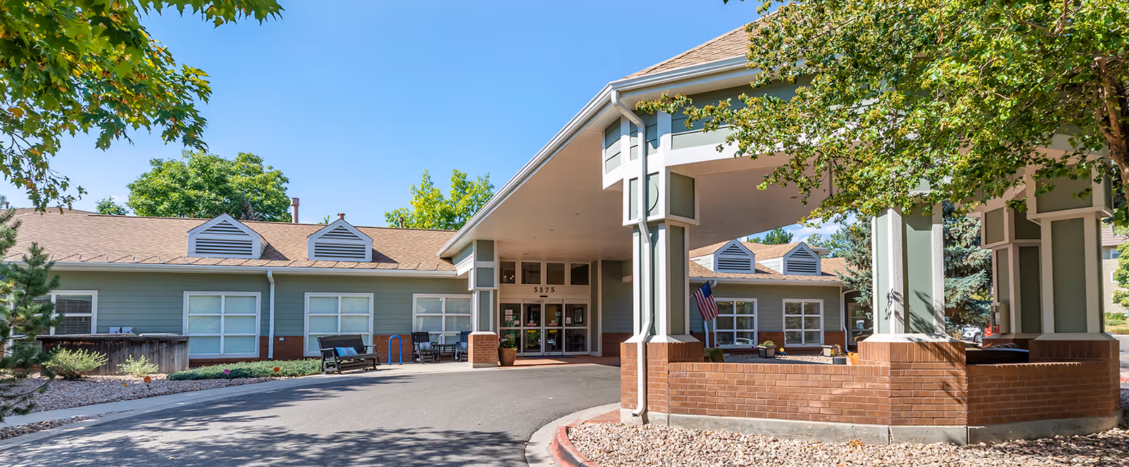 Exterior view of Brookdale Boulder Creek senior living facility showing the entrance with a covered driveway, benches, trees, and clear blue sky.