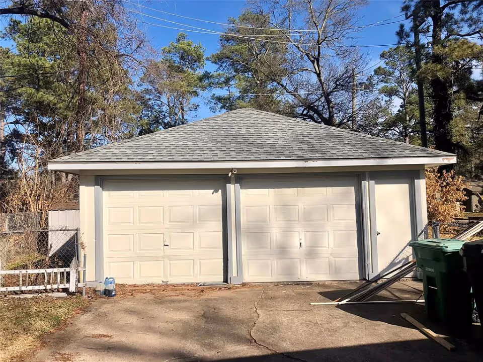 Detached two-car garage with light-colored double doors and a shingled hip roof at the end of a driveway, surrounded by trees.