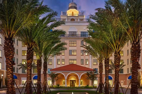 Illuminated multi-story building entrance framed by rows of palm trees and a central fountain at dusk.