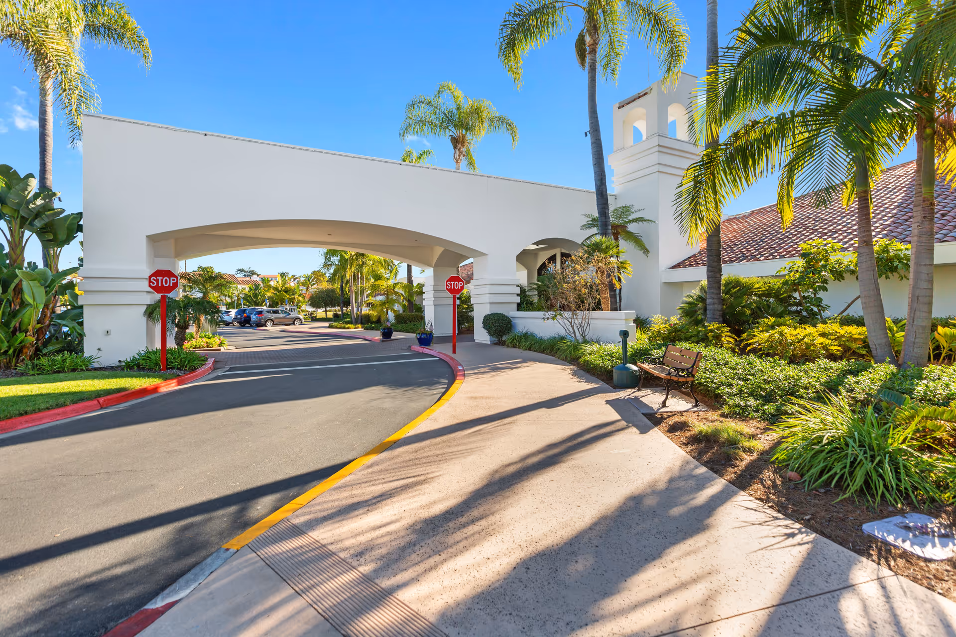 Entrance to Ocean Hills Country Club featuring a white archway with two stop signs, surrounded by palm trees and lush greenery under a clear blue sky. A bench is placed on the right side along the sidewalk.