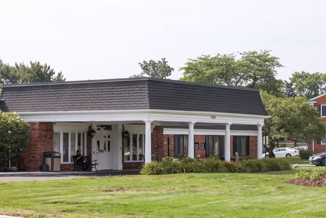 Front entrance of a single-story brick senior living facility with white columns, a covered portico and a grassy lawn.