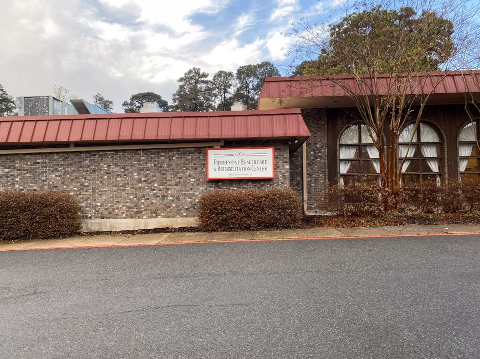 Front exterior of a brick healthcare building with a red metal roof, arched windows, shrubs and a sign reading 'Pierremont Healthcare & Rehabilitation Center'.