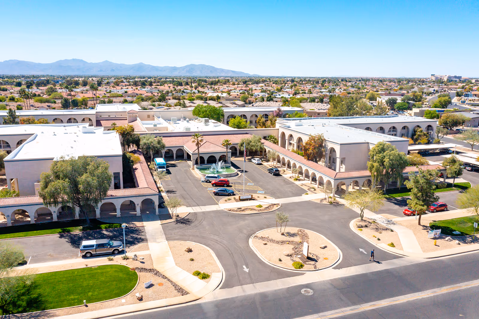 Aerial view of a senior living campus with a circular driveway and fountain, arched two-story buildings, parking areas, and surrounding neighborhood.