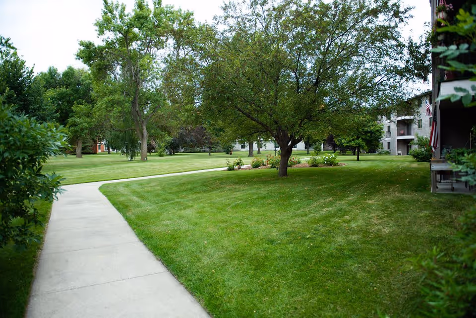 A well-maintained outdoor area with a concrete pathway curving through green grass and trees. Residential buildings are visible in the background, with balconies and an American flag displayed on one side.