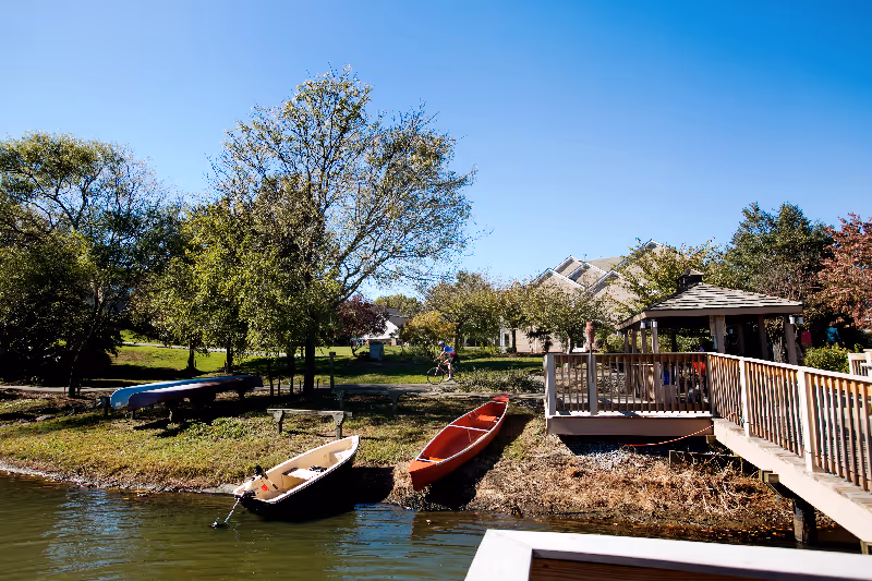Lakeside scene with a wooden dock and gazebo, several canoes along the shore, trees and houses under a clear blue sky.