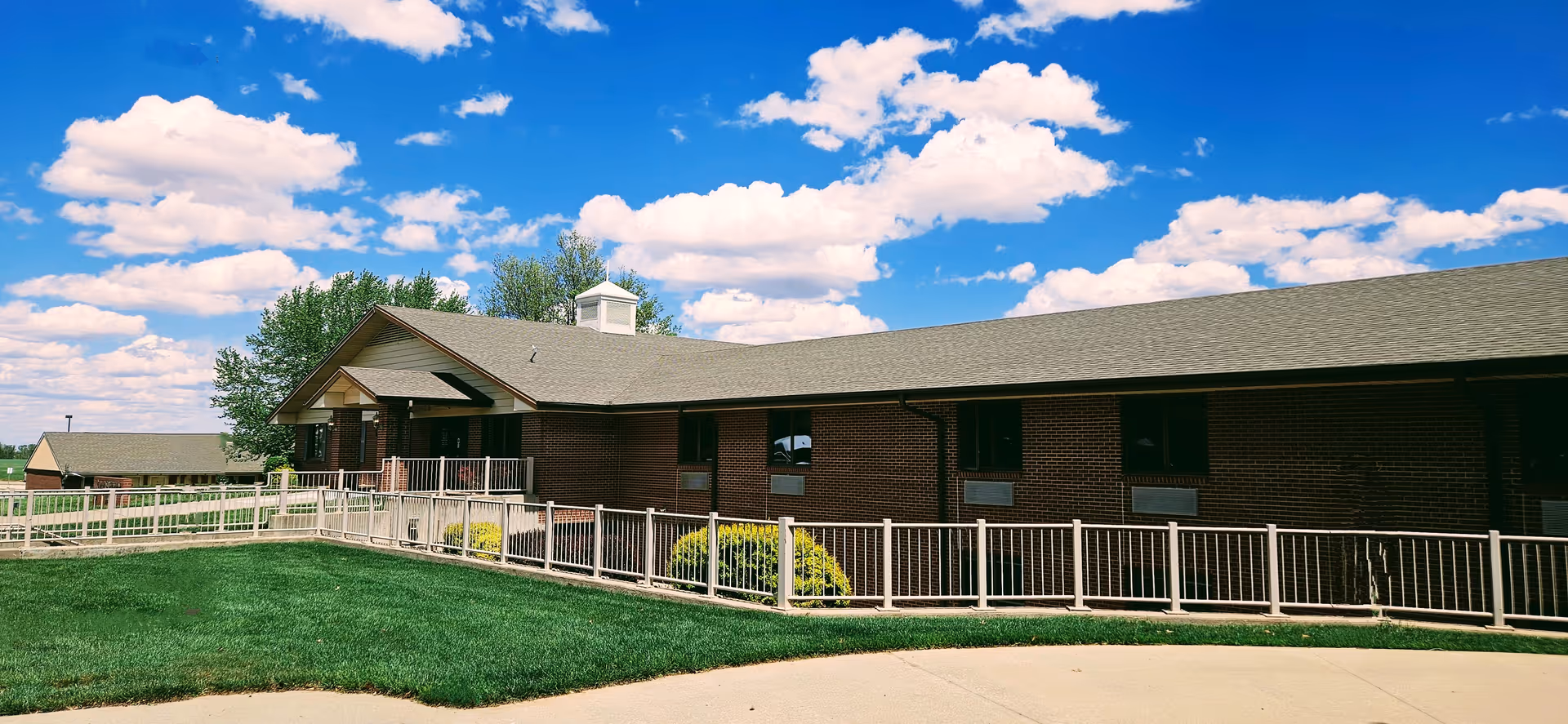 Brick senior living facility with a ramp, railing, green lawn, and blue sky above.