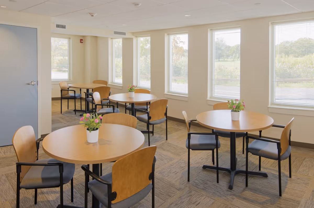 Bright dining area in a senior living facility with round tables, chairs, and potted plants.
