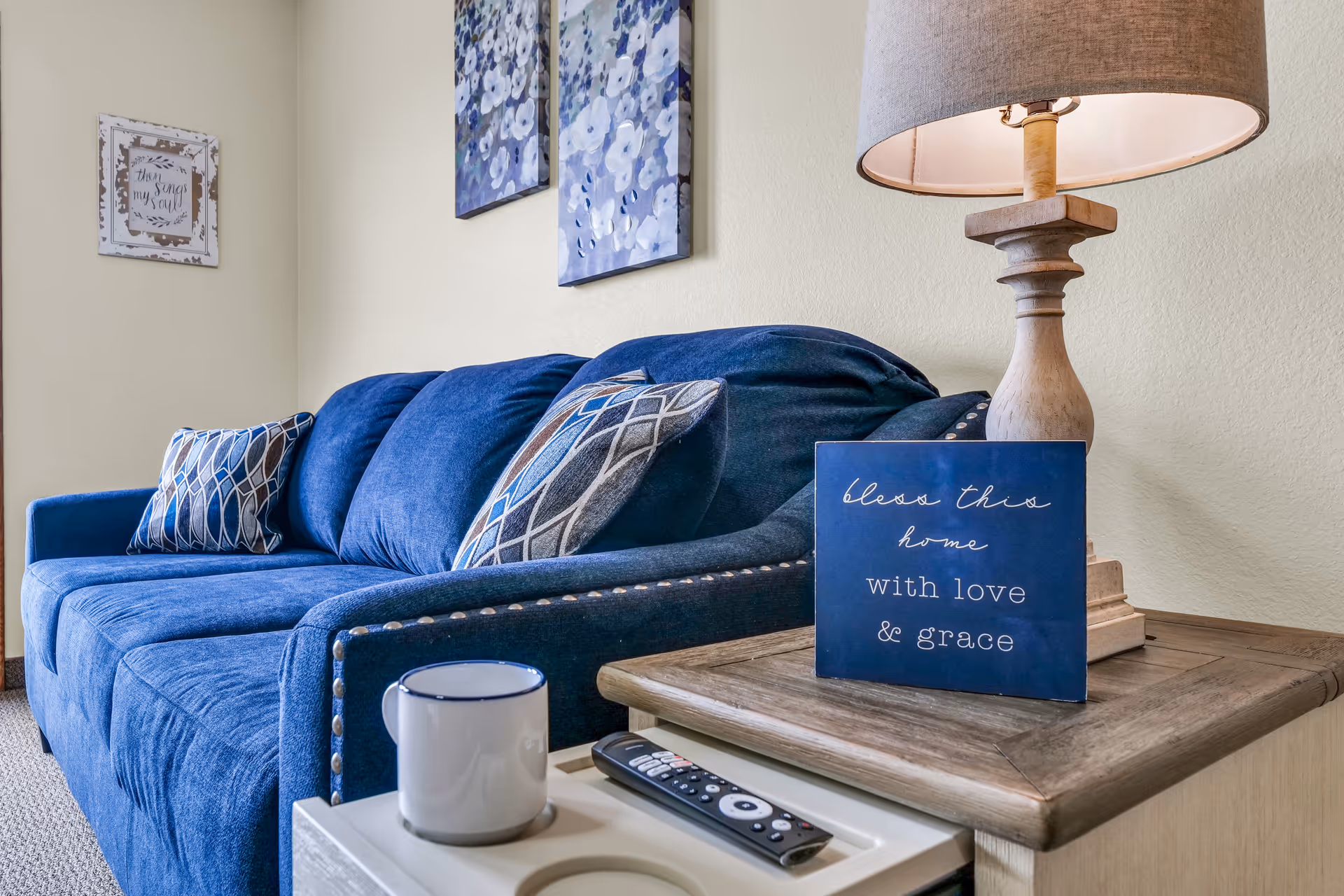 A cozy living room area featuring a blue upholstered sofa with patterned throw pillows. Next to the sofa is a wooden side table with a beige lamp, a blue decorative sign that reads 'bless this home with love & grace,' a white mug, and a TV remote control. The wall behind the sofa has two floral paintings and a framed quote.