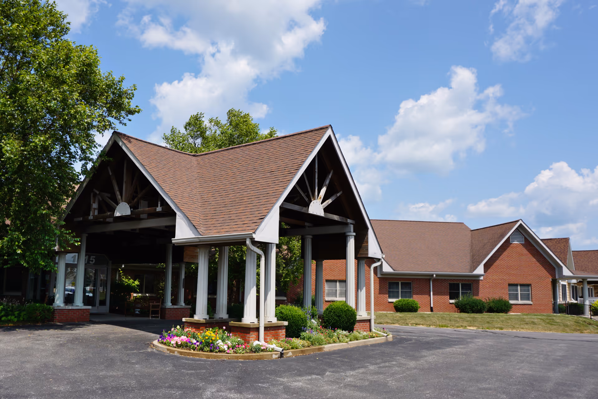 Covered entrance canopy with columns at the front of a red-brick assisted living building, a flowerbed and driveway under a blue sky.